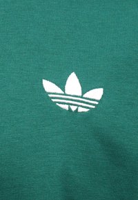 Πράσινο βαμβακερό ύφασμα με λευκό κεντημένο λογότυπο τριφυλλιού Adidas και τρεις οριζόντιες ρίγες από κάτω. Ανάγλυφο, αναπνεύσιμο υλικό.