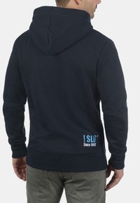 Mørkeblå hætte sweatshirt lavet af bomuldsblanding; har en kengurulomme og blå logo detalje nederst til venstre på ryggen.