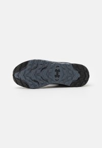 Semelle de chaussure en caoutchouc gris avec un design de bande de roulement à motifs. Présente des rainures multidirectionnelles et un logo au centre. Bordures noires texturées.