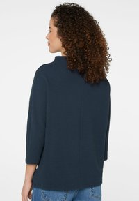 Dunkelblaues, strukturiertes Oberteil mit hohem Kragen und Dreiviertelärmeln. Der Rücken hat eine gerade Naht und einen leicht kastigen Schnitt.