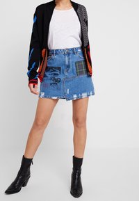 Kvinna som bär en lapptäcke denim mini kjol, vit t-shirt, svart mönstrad kofta och svarta klackade ankelstövlar mot en enfärgad bakgrund.