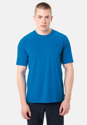 Giovane uomo con capelli corti che indossa una maglietta sportiva a maniche corte blu e pantaloni scuri, in piedi contro uno sfondo chiaro.