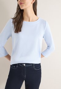 Femme portant un haut côtelé bleu clair à manches 3/4 et un jean bleu foncé avec des coutures blanches, debout contre un fond clair uni.