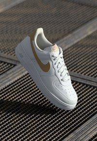 Weiße Ledersneaker mit einem goldenen Swoosh, runder Zehenpartie, perforiertem Design und weißen Schnürsenkeln. Die Sohle ist flach und strukturiert.