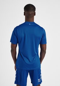 Blå atletisk trøje, korte ærmer, glat tekstur, rund hals, med hvide detaljer på skuldrene og logo på den nederste side. Shorts i matchende farve.