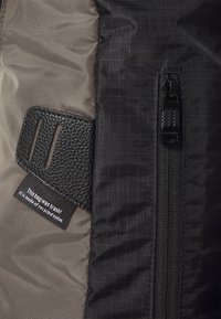 Sac en nylon recyclé avec un accent noir texturé et une fermeture éclair, doté d'un corps gris lisse et d'une étiquette visible indiquant son matériau.