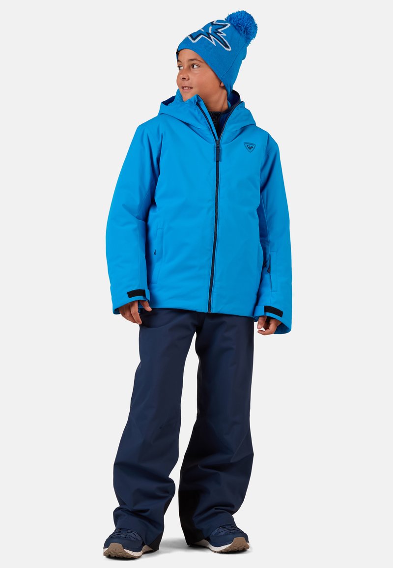 Modrá voděodolná bunda s kapucí, s logem na hrudi, spárovaná s námořnickými kalhotami a modrou pletenou čepicí s bambulí.