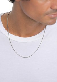 Sidabrinė grandinėlė-ant kaklo ant paprasto balto marškinėlio. Grandinėlė turi susuktą dizainą ir yra smulkiai tekstūruota, ilsisi vos virš apykaklės.