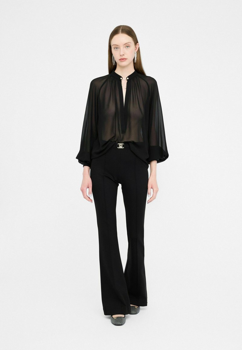 Zwarte doorschijnende blouse met hoge hals en sleutelgatdetail, gecombineerd met zwart hoge taille uitlopende broeken en gestructureerde zwarte schoenen.