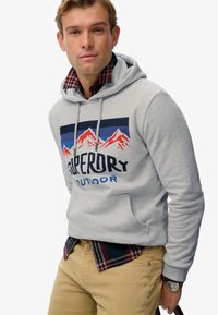 Felpa grigia con grafica di montagne, con cordini e tasca frontale, indossata sopra una camicia flanella a quadri. Sono visibili pantaloni beige e un orologio da polso.