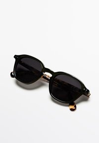 Rechthoekige zonnebril met donkere glazen, olijfgroene montuur en accenten van schildpadmotief. Voorzien van metalen details op de pootjes.