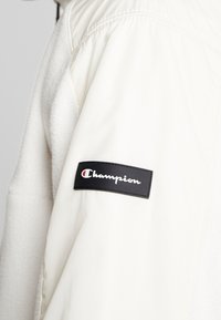 Biała rękawiczka z czarną prostokątną łatą z logo Champion w białym i czerwonym kolorze na górnej części ramienia.