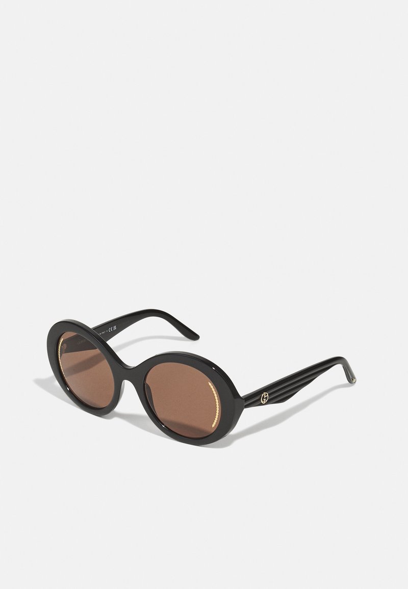 Black oval sunglasses with brown tinted lenses. Sleek plastic frame features a textured gold accent and striped arms.