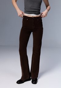 Pantalon évasé en velours côtelé marron, avec taille haute, texture subtile, design à cinq poches, porté avec des bottes à talons noires.