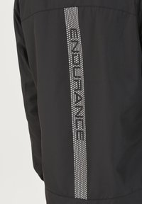 Chaqueta negra con una franja vertical reflectante que lleva el texto "ENDURANCE" en una fuente en negrita, fabricada con material suave y resistente al agua.