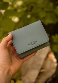 Green leather wallet with a snap closure, featuring a smooth texture and embossed logo on the front. Handheld against a blurred green background.
