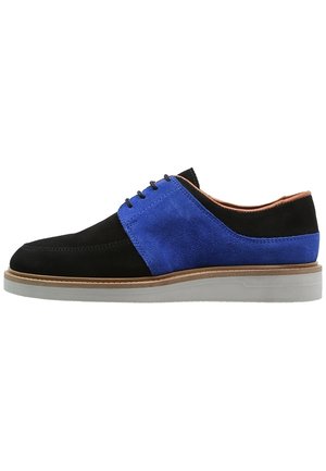 Zapato oxford de hombre en ante negro y azul con cordones negros, ribete marrón claro y suela de goma blanca, visto de lado sobre un fondo blanco.
