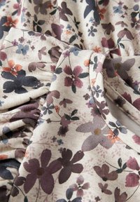 Blommönstrad tyg i beige, med lila, orange och blå blommor. Mjuk textur med rynkdetaljer och ribbade accenter.