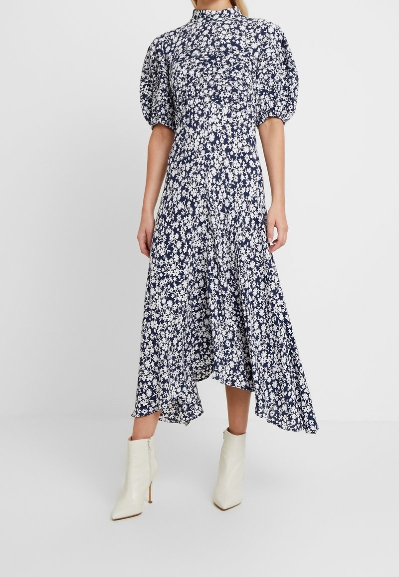 Femme portant une robe midi bleu marine à imprimé floral blanc avec manches bouffantes, associée à des bottines blanches à bout pointu.