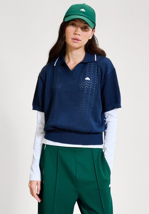 Vrouw met een groene pet, marineblauw kort mouw gebreid poloshirt over een witte lange mouw shirt, en groene broek met zijzakken, staand tegen een effen achtergrond.