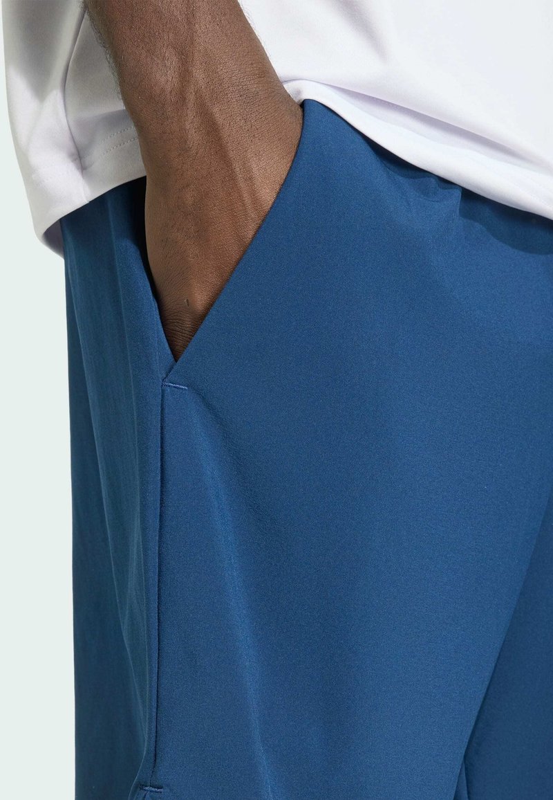 Hand in der Tasche einer blauen Hose, getragen mit einem weißen Hemd, wobei der Fokus auf der Stoffstruktur und den Details der Taschennaht liegt.