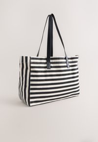 Borsa tote rettangolare a righe bianche e nere con spallacci neri, in piedi su uno sfondo chiaro e semplice.