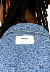 Denimjack met een lichtblauwe kleur, voorzien van een hartpatroon en een wit etiket met "SIXTH JUNE PARIS" gestikt op de achterkant.