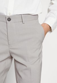 Pantaloni grigi chiari in un tessuto liscio con vestibilità slim, dotati di chiusura con bottone e tasche laterali, abbinati a una camicia bianca.