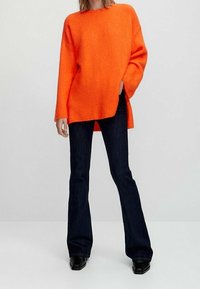 Pull orange surdimensionné avec un col rond, fentes sur les côtés et épaules tombantes, associé à un jean noir évasé et des bottines noires.