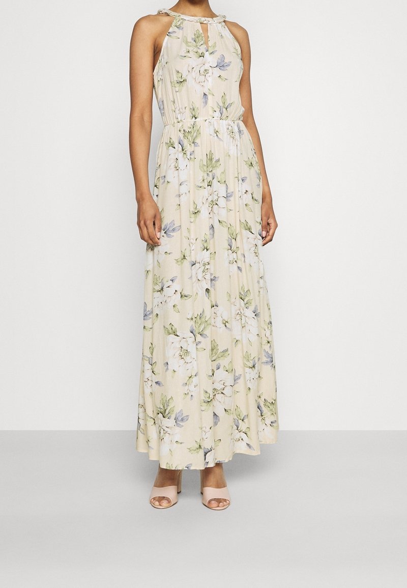 Femme portant une robe beige sans manches, longue jusqu'au sol, ornée d'un imprimé floral vert et blanc, associée à des talons ouverts rose clair.