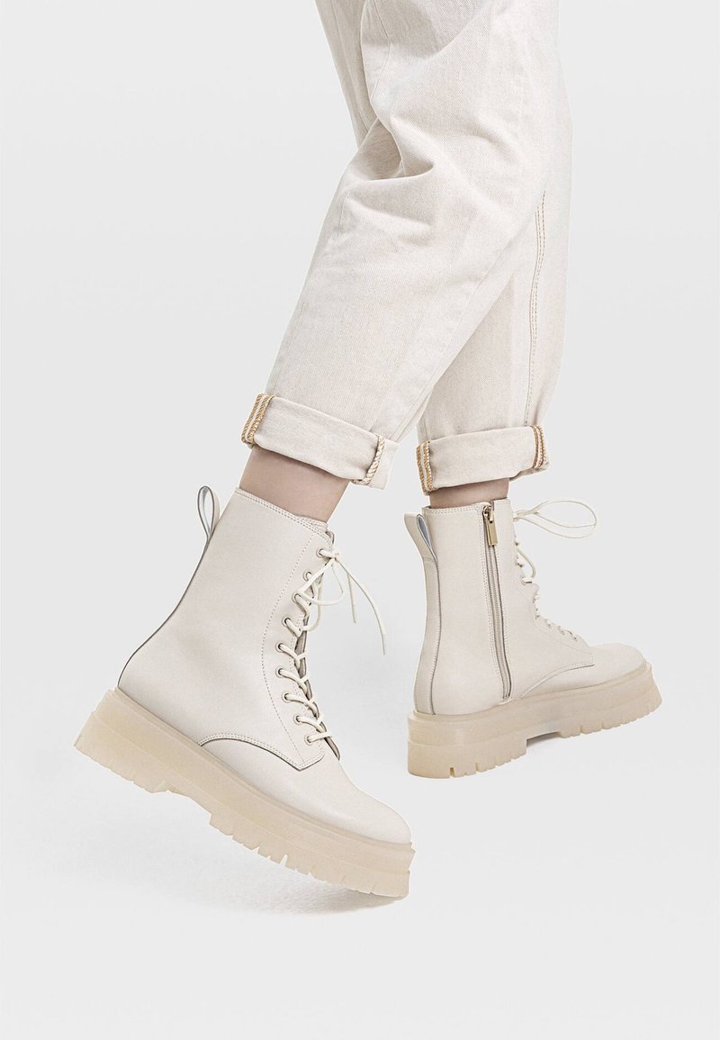 Personne portant des bottes beige clair à lacets avec des fermetures éclair latérales et un pantalon retroussé sur un fond uni.