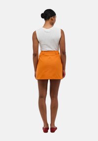 Jupe en coton orange avec une taille cintrée et deux poches arrière. Portée avec un top sans manches blanc texturé et des chaussures rouges.