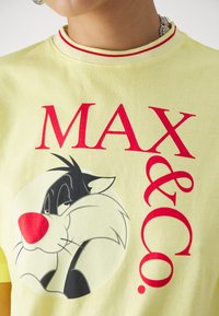 Camiseta de algodón amarilla con un cuello redondo bordeado en rojo. Presenta un gráfico de Silvestre el Gato y el texto "MAX & CO." en rojo negrita.