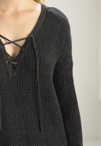 Schwarzer Strickpullover mit V-Ausschnitt-Design, der ein Schnürdetail am Ausschnitt aufweist. Breite Ärmel und eine strukturierte Oberfläche verleihen einen gemütlichen Look.