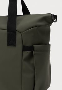 Mørkegrønn toteveske med svarte håndtak, frontlomme og en løkke for feste på en lys, ensfarget bakgrunn.