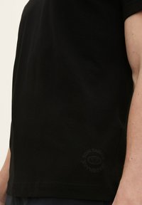 Svart bomulls-T-skjorte med korte ermer, rund hals og en diskret logo trykt på nedre side. Glatt tekstur.