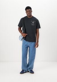 Black cotton t-shirt with graphic print, blue wide-leg denim jeans, and navy sneakers featuring three white stripes. Small crossbody bag.