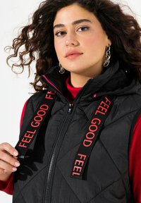 Svart vadderad väst med huva, prydd med röda "FEEL GOOD" remmar och en framdragkedja. Bärs över en röd polotröja, vilket framhäver texturerade detaljer.