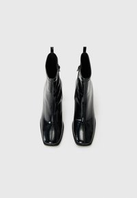 Bottines noires en cuir verni avec un embout carré, texture lisse, fermeture éclair latérale et silhouette élancée avec un talon légèrement rehaussé.
