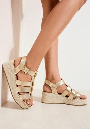 Jalad, mis kannavad kuldseid kiilkontsaga sandaale, millel on kolm rihma ja üks pahkluu pandlaga rihm, seisavad heledal põrandal valge seina taustal.