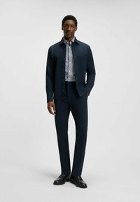 Laivasto sininen nappikiinnitteinen takki, jossa on rintatasku, musta-valkoisen ruudullisen paidan päällä, yhdistettynä räätälöityihin laivasto sinisiin housuihin ja mustiin kenkiin.