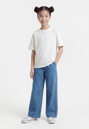 Fată tânără cu coafură cu două cocuri, purtând un tricou alb, blugi evazați albaștri și adidași albi, stând cu o mână în buzunar.