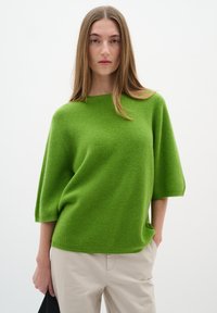 Green knitted sweater with a relaxed fit, wide sleeves, and a ribbed texture. Paired with light beige pants. Simple design, minimal accents.
