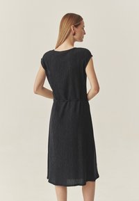 Crna haljina do koljena s opuštenim krojem, sa kratkim rukavima i suženim strukom. Teksturirana tkanina s suptilnim vertikalnim linijama.