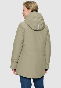 Vrouw met kort blond haar, gekleed in een lichtbeige parka met capuchon en donkerblauwe spijkerbroek, met haar rug naar de camera gekeerd tegen een effen achtergrond.