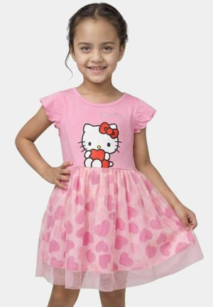 Fille souriante en robe rose avec Hello Kitty tenant un cœur rouge en haut et une jupe en tulle avec des cœurs rose foncé.