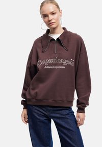 Sudadera marrón con cuello alto, cierre de media cremallera y texto en blanco "Departamento Atlético de Copenhague". Corte relajado, textura suave.