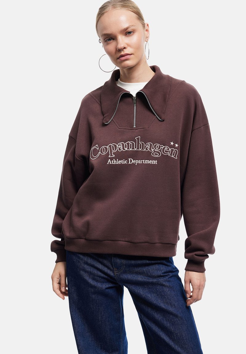 Sudadera marrón con cuello alto, cierre de media cremallera y texto en blanco "Departamento Atlético de Copenhague". Corte relajado, textura suave.