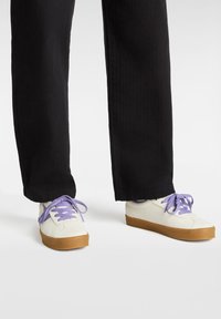 White sneakers with purple laces, featuring a textured upper and brown rubber soles, paired with black, straight-leg pants.