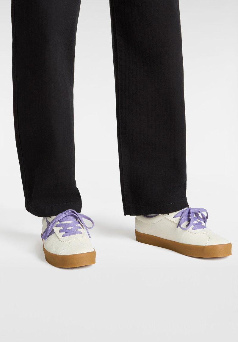 White sneakers with purple laces, featuring a textured upper and brown rubber soles, paired with black, straight-leg pants.